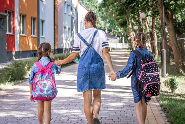 Young mom is accompanied little girls to school, back view.