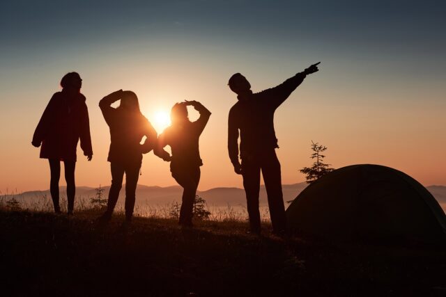 A silhouette of group people have fun at the top of the mountain near the tent during the sunset