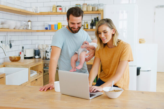 Busy Family In Kitchen At Breakfast With Parents Working On Laptop And Caring For Baby Son